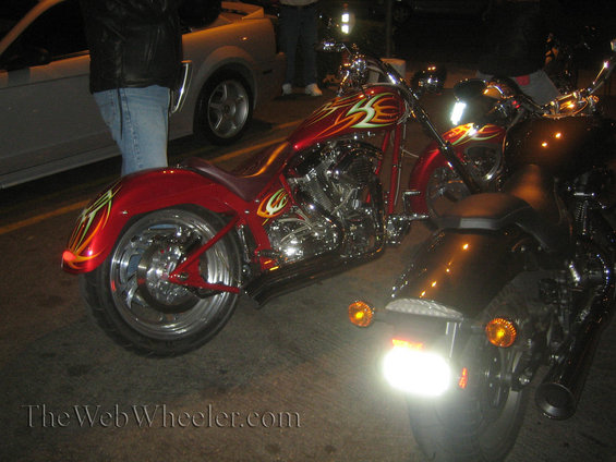 Two custom motorcycles and a silver car parked at night. The motorcycles have flame graphics