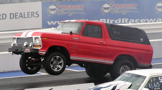Red 1979 Ford Bronco with white racing stripes on hood and side, front wheels off the ground at a drag strip