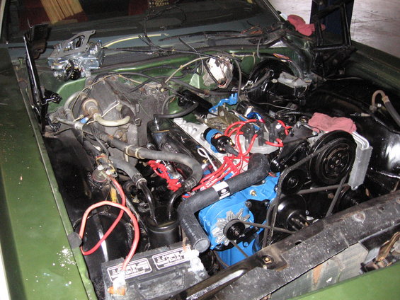 Engine bay view showing a blue engine block, carburetor, battery, and various hoses and wires
