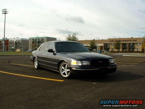 Dark sedan parked on asphalt with yellow lines. Building in background has text 'mtl_cv@crownvic.net'