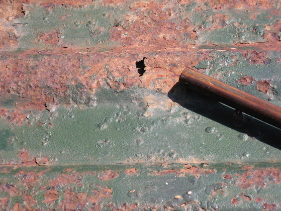 Corrugated metal surface with extensive rust and peeling green paint. A cylindrical metal object casts a shadow