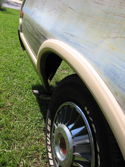 Vehicle's rear quarter panel with woodgrain-like paint and a beige fender flare over a wheel with a chrome hubcap and white-lettered tire
