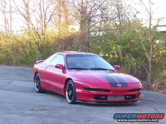 Red 1997 Ford Probe with black racing stripes on the hood, parked on a paved surface with vegetation in the background