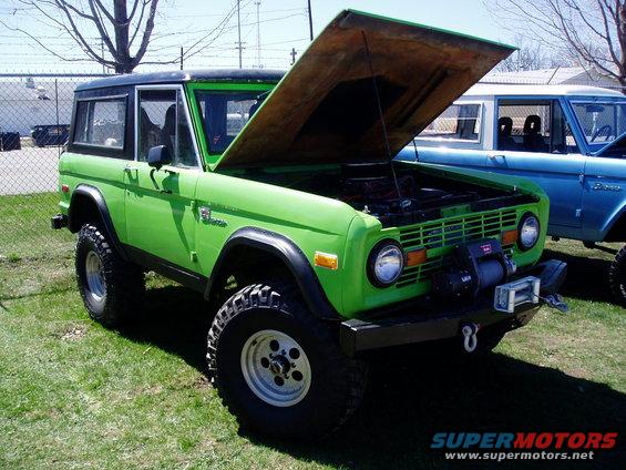 Green Ford Bronco with hood open, black top, and winch on front bumper. Blue Ford Bronco parked behind it