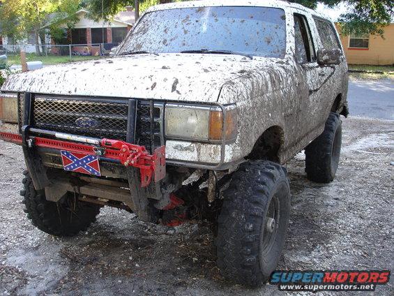 1989 Ford Bronco covered in mud, with a Hi-Lift jack and Confederate flag on the front bumper