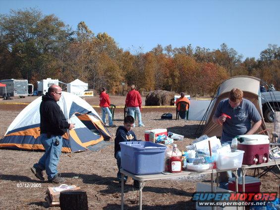 People gathered outdoors near tents with a table of food and coolers. A date stamp reads 03/12/2005