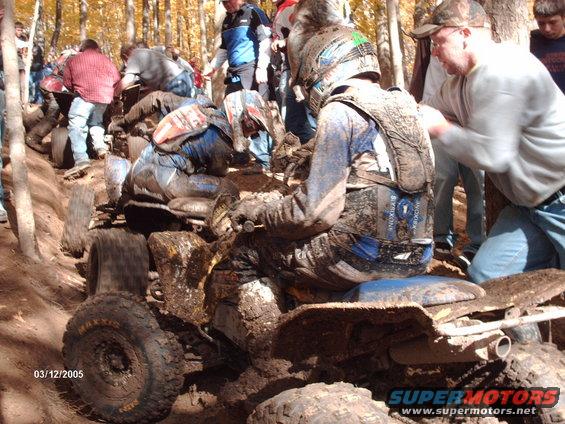 Quad bikes stuck in mud with riders wearing protective gear, spectators in background