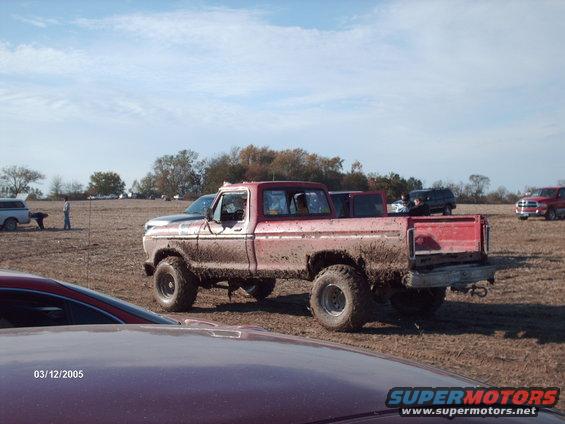Mud-covered red pickup truck with large tires parked in a field, other vehicles and people visible in the background