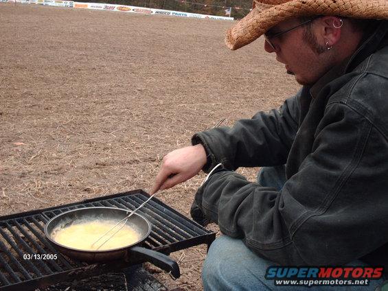 guy in cowboy hat cooking eggs in a pan on a grill
