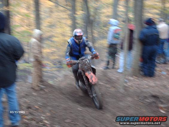 Dirt bike rider wearing blue and orange gear on a muddy track in the woods