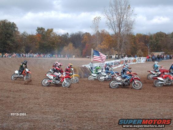 Motocross race with multiple dirt bikes on a dirt track, riders in gear, American flag, spectators in background