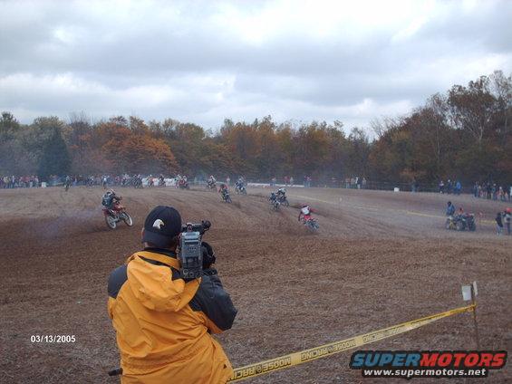 Motocross race in progress with multiple riders on a dirt track, spectators in the background, and a cameraman in the foreground. Date stamp reads 03/13/2005