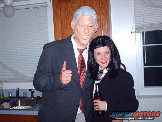 Guy in a Bill Clinton mask gives a thumbs up next to a woman in a Hillary Clinton wig and suit, holding a beer