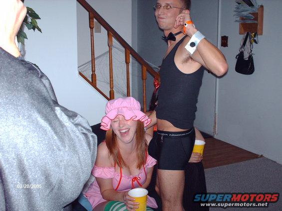 Two people in costumes, one in a pink dress and bonnet, the other in a tank top and underwear, holding cups at a party