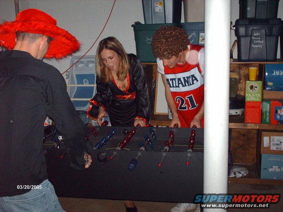 Three people in costumes play foosball in a basement. One wears a large red hat, another a black robe, and the third a basketball jersey