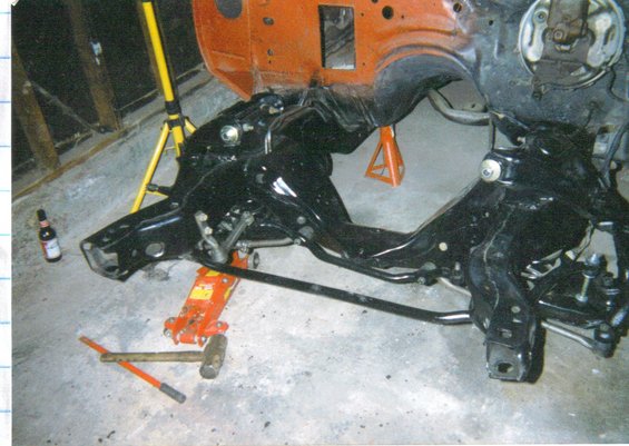 Car frame and suspension components on jack stands and a hydraulic jack, with tools and a beer bottle nearby