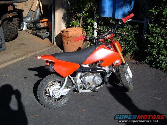 Orange dirt bike parked outside a garage. It has a black seat and handlebars