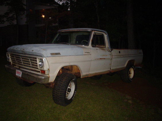 White 1967 Ford F-250 pickup truck with a rusty bed, lifted suspension, and off-road tires, parked on grass