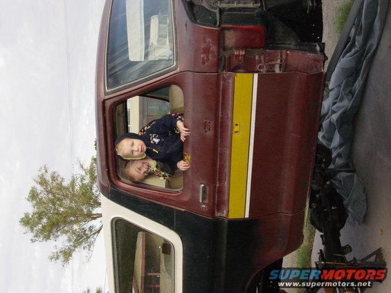 Two children look out the driver's side window of a maroon and white SUV, possibly a Ford Bronco. The vehicle appears to be undergoing repairs or is partially disassembled