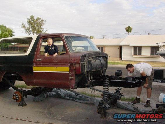 Kid in driver's seat of a maroon and black Ford Bronco body being worked on by a man. The chassis and axles are removed