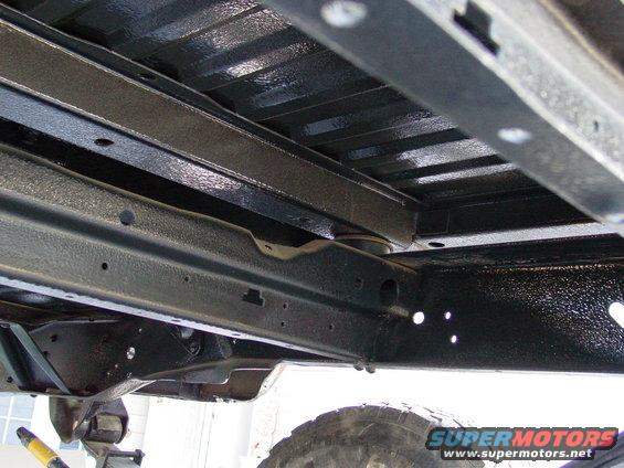 underbody of a vehicle with frame rails and crossmembers coated in black protective material, tire visible at bottom right
