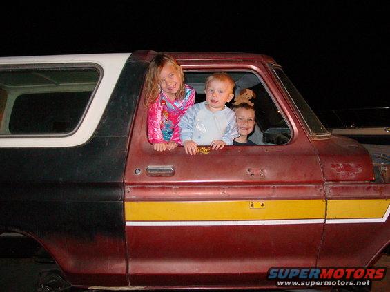 Three children look out the windows of a red and black SUV with a yellow stripe