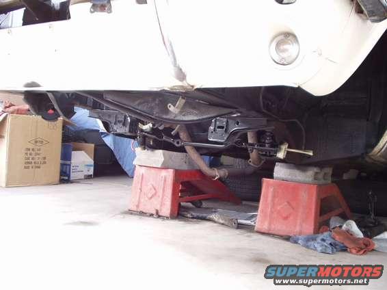 Underside of a vehicle on jack stands, showing suspension and exhaust components. Cardboard boxes are nearby