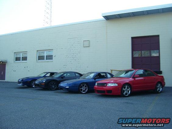 Four cars parked in a row outside a building. The cars are a blue Camaro, a black coupe, a blue coupe, and a red sedan