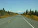 1982 Chevrolet C/K Pickup - A highway stretches towards snow-capped mountains, flanked by autumn foliage and evergreen trees under a clear blue sky.