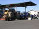 1982 Chevrolet C/K Pickup - A blue pickup truck tows a large tractor on a trailer past gas pumps under a covered canopy.