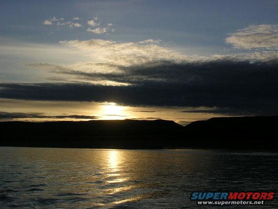 sun setting behind hills over water, clouds in sky