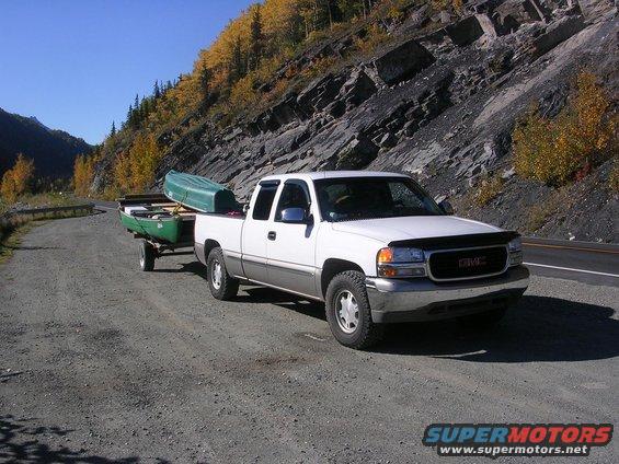 White GMC pickup truck towing a trailer with a green canoe on a gravel road