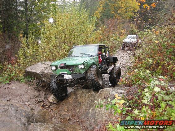 Green off-road vehicle with large tires navigating a rocky trail, another white vehicle behind it