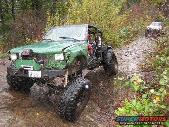 Green off-road vehicle with a roll cage and large tires navigating a rocky stream bed