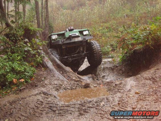 Green off-road vehicle with large tires is tilted on a muddy, wooded trail, with one wheel in a ditch