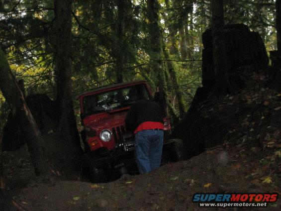 Red Jeep Wrangler off-road in a forest, front-left quarter view, with a person standing beside it