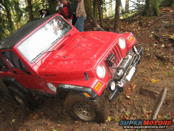 Red Jeep Wrangler with winch stuck in muddy terrain, people watching in background