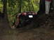 1990 Ford Bronco - A red Jeep Wrangler tackles a challenging muddy off-road course surrounded by dense forest.
