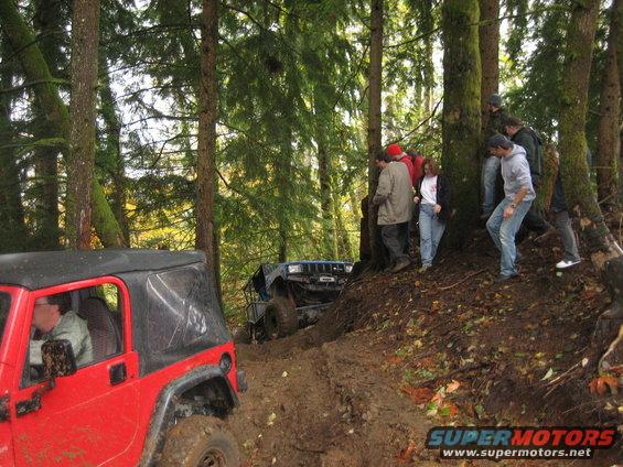 Red Jeep Wrangler and blue off-road vehicle on muddy trail with people watching