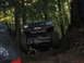 1990 Ford Bronco - An off-road SUV navigates a steep, muddy incline in a wooded area, with another vehicle partially visible to the left.