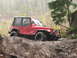 1990 Ford Bronco - A red Jeep Wrangler navigates a challenging muddy off-road trail in a wooded area.