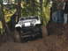 1990 Ford Bronco - A white Jeep Wrangler tackles a challenging muddy trail with onlookers observing.