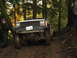 1990 Ford Bronco - A Jeep Cherokee is shown off-roading on a muddy trail surrounded by trees.