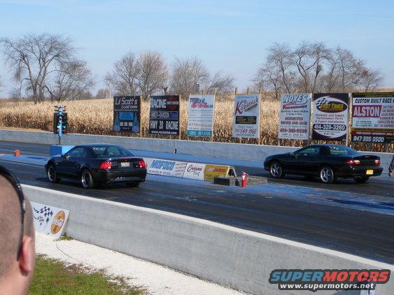 Two black muscle cars racing on a drag strip with sponsor banners in the background