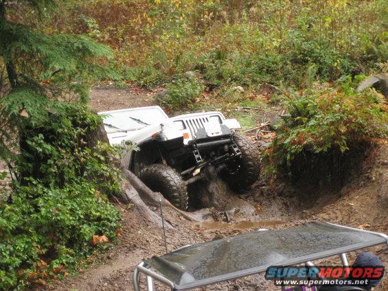 White Jeep Wrangler stuck in mud on a trail, another vehicle's roof visible in foreground