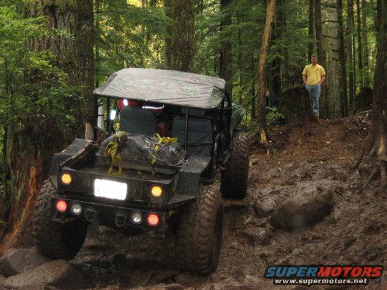 Off-road vehicle with a camo tarp roof driving on a rocky, muddy trail in a forest. A man stands on a hill in the background