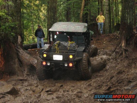 Black off-road vehicle with a camouflage top navigates a muddy forest trail