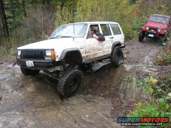 White Jeep Cherokee on rocky terrain, driver visible, mud-splattered
