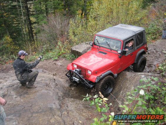Red Jeep Wrangler navigating a rocky off-road trail with a person in the driver's seat and another person crouching nearby