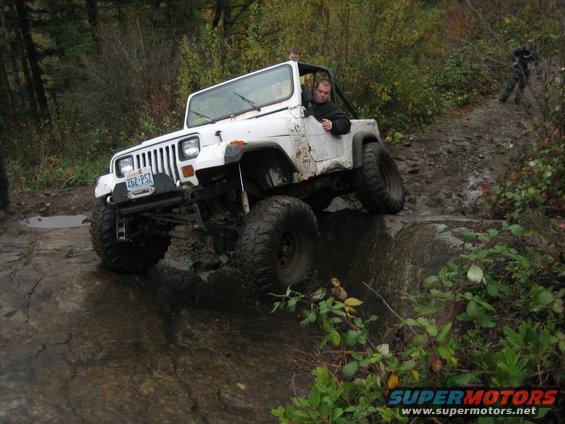 White Jeep Wrangler driving over rocks on a muddy trail. Driver is visible. Another person is in the background
